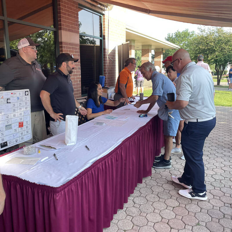 People gather around a table with a red table skirt outside a building under a beige awning on a sunny day. The table has papers and other items on it, likely for the 2023 Golf Outing. One person is holding a pen, while others interact with the individuals behind the table.