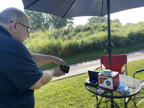 A person taking a photo with a smartphone outdoors on a sunny day during a 2023 golf outing. They're next to a table with a large umbrella, which has various items, including a box, laptop, wireless charger, and container. A red folding chair is also visible nearby.