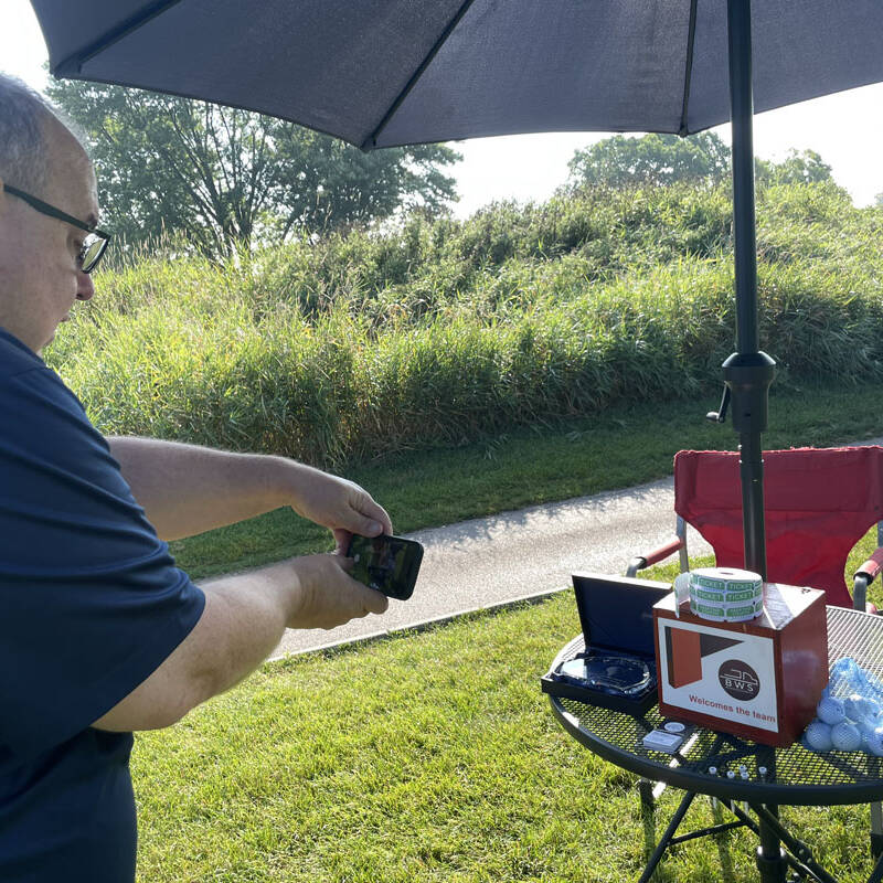 A person taking a photo with a smartphone outdoors on a sunny day during a 2023 golf outing. They're next to a table with a large umbrella, which has various items, including a box, laptop, wireless charger, and container. A red folding chair is also visible nearby.