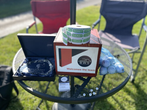 A small round table holds a "BWS Welcomes the Team" sign, two rolls of raffle tickets, a plaque, business cards, a pile of small items, and a mesh bag of golf balls. Foldable chairs and a grassy area set the scene for the 2023 outing.