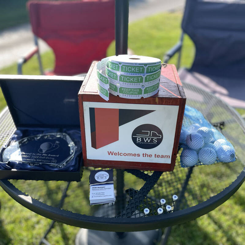 A small round table holds a "BWS Welcomes the Team" sign, two rolls of raffle tickets, a plaque, business cards, a pile of small items, and a mesh bag of golf balls. Foldable chairs and a grassy area set the scene for the 2023 outing.