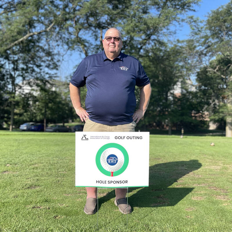 A man in a blue polo shirt and khaki shorts stands on a golf course holding a sign that reads "2023 Golf Outing" and "Hole Sponsor." The background features trees and a sunny sky.