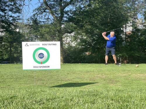 A man in a blue shirt and shorts is swinging a golf club at a 2023 golf outing. A sponsorship sign for the Penn State Abington Athletics Golf Outing is visible in the foreground, indicating a hole sponsor. Trees and greenery surround the course.