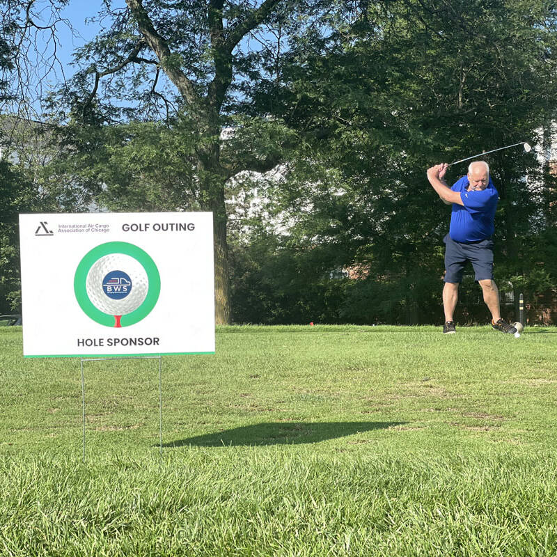 A man in a blue shirt and shorts is swinging a golf club at a 2023 golf outing. A sponsorship sign for the Penn State Abington Athletics Golf Outing is visible in the foreground, indicating a hole sponsor. Trees and greenery surround the course.