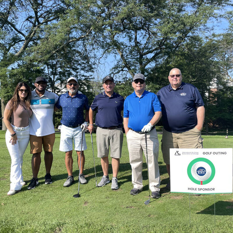 A group of six people stands on a golf course, smiling at the camera during the 2023 Golf Outing. They are dressed in casual golf attire and holding golf clubs. A sign next to them reads "Golf Outing Hole Sponsor." Trees and grass surround them on a sunny day.