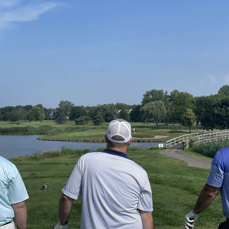 Three men stand on a golf course during the 2023 Golf Outing, facing a lake with a green on the other side. They wear golf attire and hats, with one man in a light blue shirt, another in white, and the third in dark blue and purple. Trees and buildings dot the distant landscape.