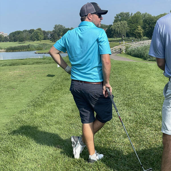 A man in a blue shirt and dark shorts stands on a grassy area next to a golf course during a 2023 outing, holding a golf club. He is wearing a baseball cap and sunglasses, looking off into the distance. A small lake and a path surrounded by trees are visible in the background.