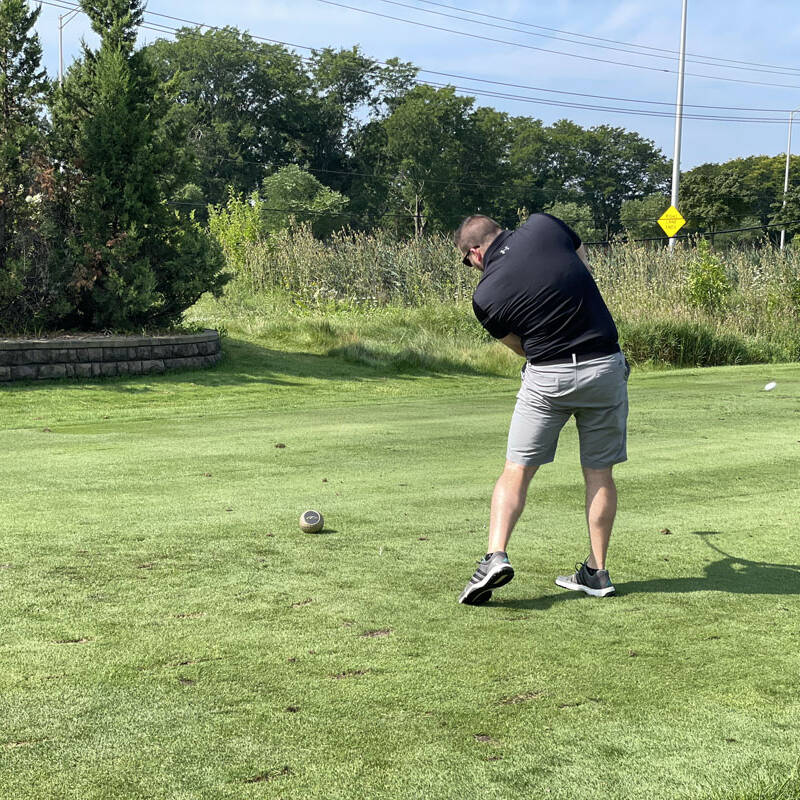 A man in a black shirt and gray shorts is seen from the back, swinging a golf club on a green golf course during a 2023 outing. There are trees and a road with a yellow sign in the background. It's a sunny day.