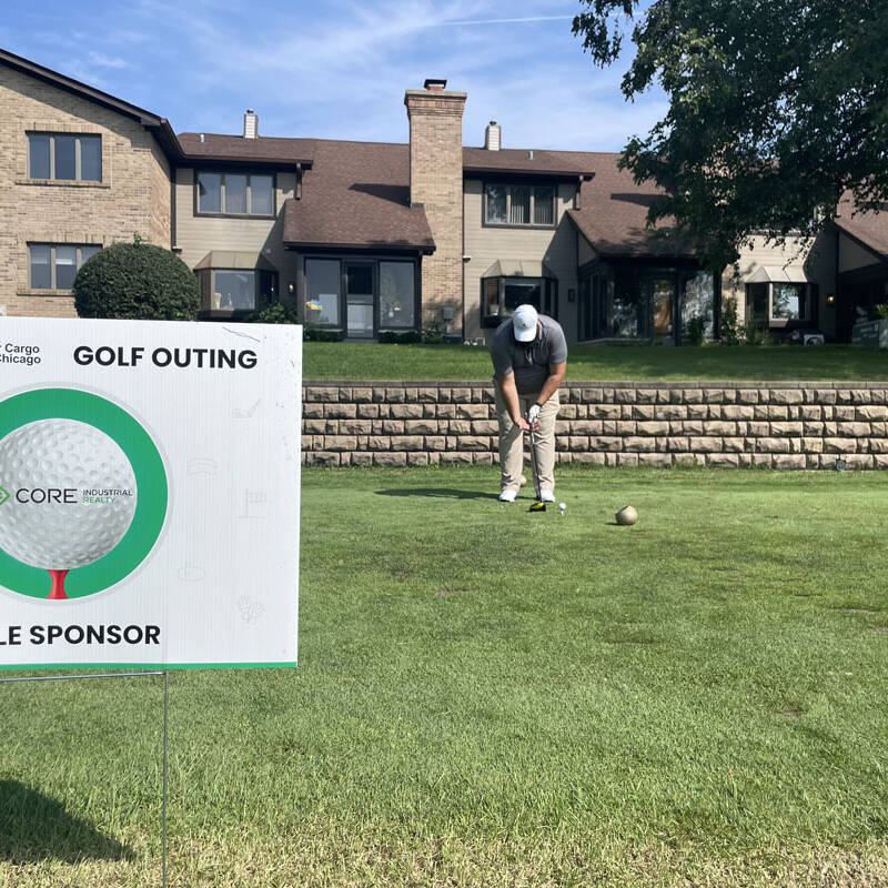 A golfer prepares to putt on a green in front of a residential building. A sign in the foreground reads, "International Air Cargo Association of Chicago: 2023 Golf Outing, Core: Hole Sponsor." Trees and a clear, blue sky are visible in the background.