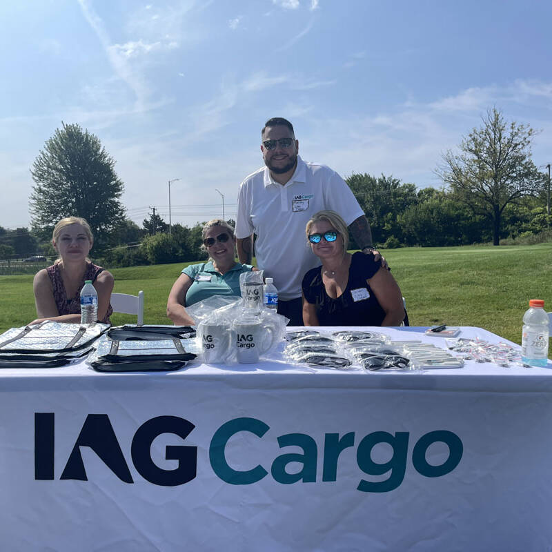 Four people are sitting and standing behind a table covered with a white cloth displaying the logo "IAG Cargo." The table is set outside on a sunny day during the 2023 golf outing, with a green field and trees in the background. Promotional items, including bags and bottles, adorn the table.