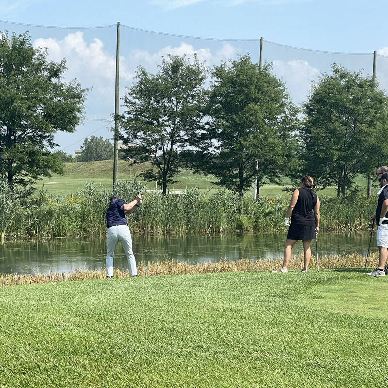 Three people are on a golf course near a water hazard during their 2023 golf outing. One person is attempting to retrieve a golf ball from the water while the other two are standing nearby watching. Trees and protective netting are visible in the background under a blue sky.