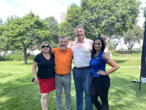 Four people standing on a grassy area in front of trees, posing for a group photo on a sunny day during the 2023 Golf Event. They are dressed casually and smiling at the camera.