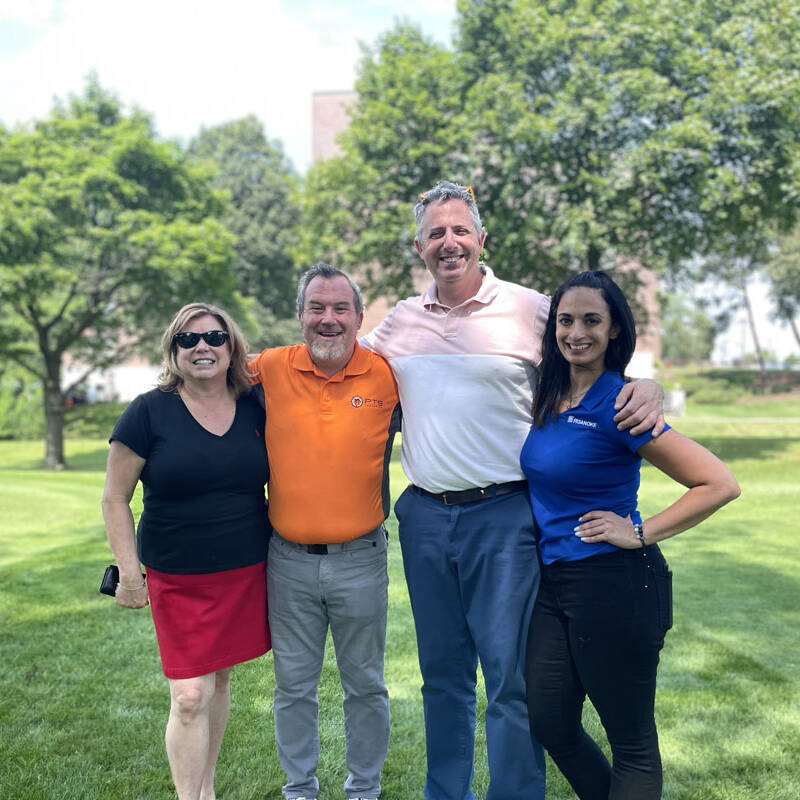 Four people standing on a grassy area in front of trees, posing for a group photo on a sunny day during the 2023 Golf Event. They are dressed casually and smiling at the camera.