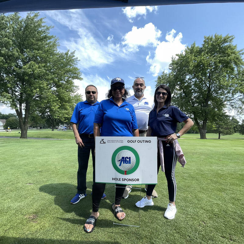 Four people stand on a golf course, posing behind a sign for the 2023 golf outing sponsored by ACI. The group is under a canopy with green grass and trees in the background. They are dressed in casual golf attire, and the weather appears sunny with a clear sky.