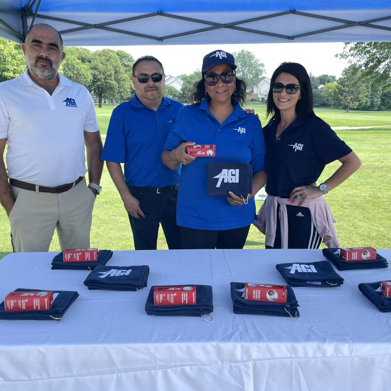 Four people are standing under a tent at the 2023 Golf Outing. They are behind a table with branded items laid out on it, all wearing clothing featuring the same logo. The background shows a grassy, open space with trees. The mood appears cheerful and relaxed.