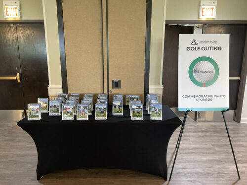 A table covered with a black cloth holds numerous framed photos. A sign next to the table reads "GOLF OUTING 2023" with an image of a golf ball and logos of the organizers. The event appears to be sponsored by Roanoke. Two closed double doors are seen in the background.