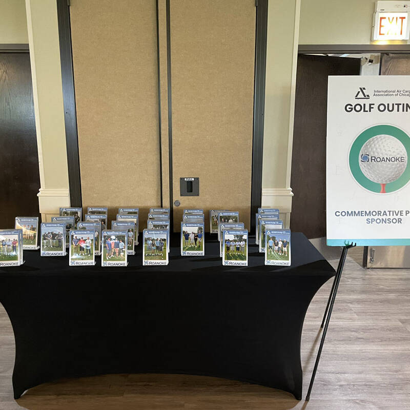 A table covered with a black cloth holds numerous framed photos. A sign next to the table reads "GOLF OUTING 2023" with an image of a golf ball and logos of the organizers. The event appears to be sponsored by Roanoke. Two closed double doors are seen in the background.