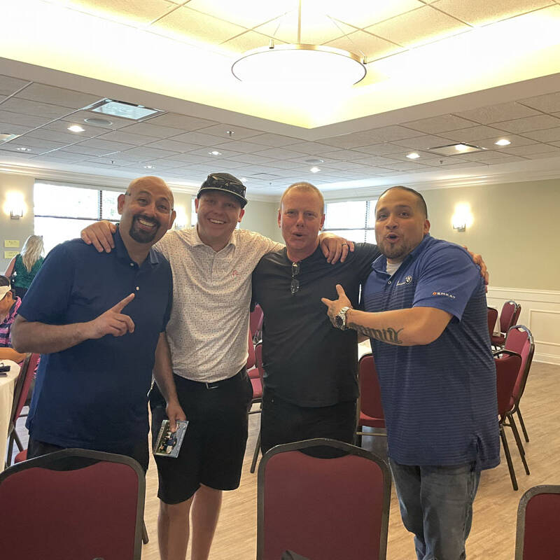 Four men are standing close together in a room with tables and chairs. The man on the left is holding up one finger, the man next to him is wearing a hat, and the other two men are smiling, likely reflecting on their golfing outing in 2023. The room has bright lighting and a relaxed atmosphere.