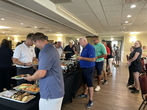 A group of people stands in line at a buffet table in a brightly lit room during the 2023 Golf Outing. Various dishes, including salad, bread, and desserts, are displayed on the table. Some individuals are serving themselves, while others wait patiently. The atmosphere appears casual and friendly.