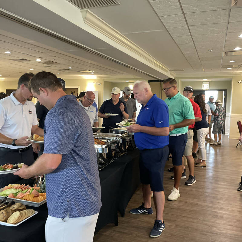 A group of people stands in line at a buffet table in a brightly lit room during the 2023 Golf Outing. Various dishes, including salad, bread, and desserts, are displayed on the table. Some individuals are serving themselves, while others wait patiently. The atmosphere appears casual and friendly.