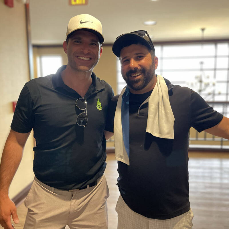 Two men stand indoors with their arms around each other, smiling at the camera. Both are wearing casual 2023 golf attire, including caps. The man on the left is dressed in a black shirt and light-colored pants, while the man on the right wears a black shirt and white shorts—a perfect snapshot of their outing.