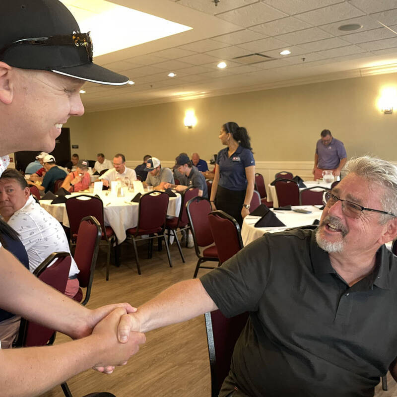 Two men are shaking hands in a banquet hall during the 2023 outing. The man on the left wears a patterned shirt and a black cap, while the man on the right, possibly discussing their recent golf game, wears glasses and a gray polo shirt. Other people in casual attire are seated at tables in the background.