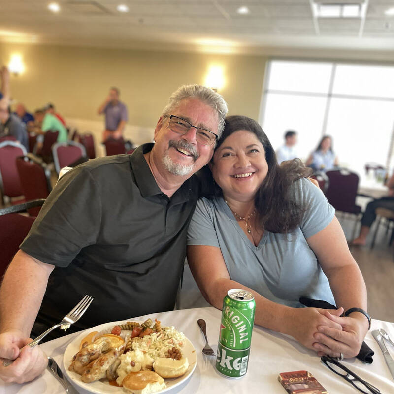 A joyous couple sits at a table in a dining area, smiling and leaning towards each other. The man holds a fork and has a plate of food in front of him, while the woman enjoys a can of A&W root beer beside her. It's clear that this 2023 outing is just what they needed after a fun day of golf. Other people dine and converse in the background.