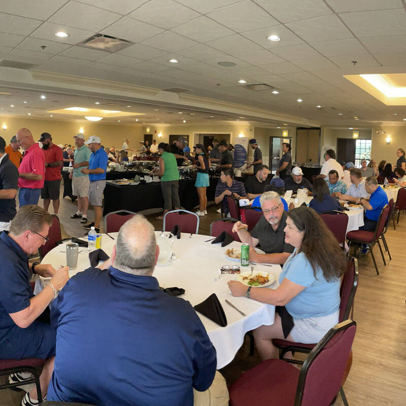 A large group of people gathers in a banquet hall with round tables and red chairs. Some are seated and eating, while others are standing and serving themselves from a buffet. The room is brightly lit with overhead lights, and large windows offer a view of the 2023 golf outing happening outside.