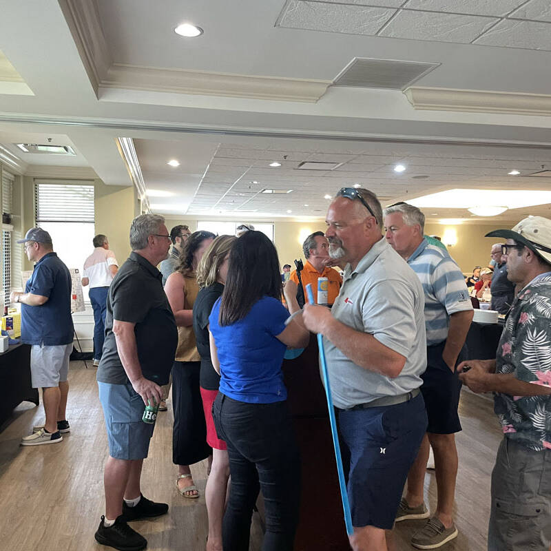 A group of people stand and converse around a high-top table in a brightly lit room with large windows. Some participants wear casual clothing, while others sport summer hats and sunglasses, evoking a relaxed outing. Tables with raffle prizes from the 2023 golf event line the wall in the background.