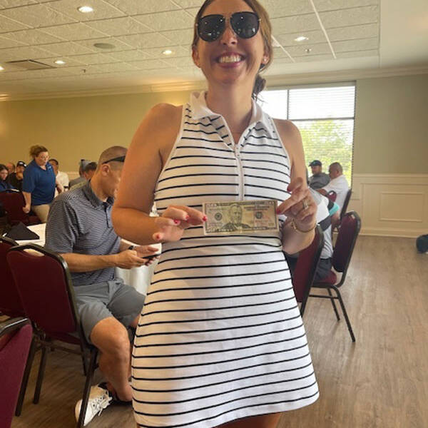 A person wearing sunglasses and a white striped dress holds a $50 bill, smiling at the camera. They are in a room with red chairs and a long table where other people are seated, possibly planning their next outing. A window with blinds is in the background, letting in natural light.