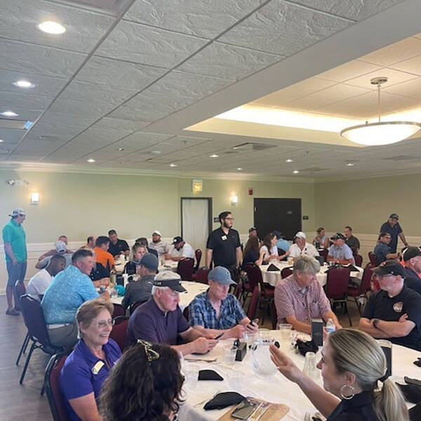 A large group of people sits and stands in a banquet hall with round tables, engaging in conversations and eating. The room, illuminated by recessed lighting from the white ceiling and natural light through large windows, buzzes with excitement about the upcoming 2024 golf outing.