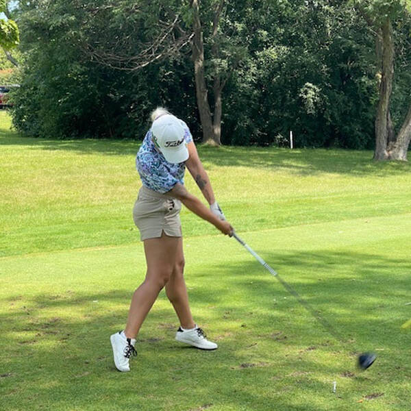 A person is captured in mid-swing on a golf course during a 2024 golf outing, wearing a white cap, patterned blue shirt, and beige shorts. The lush green fairway and trees are visible in the background on a sunny day.