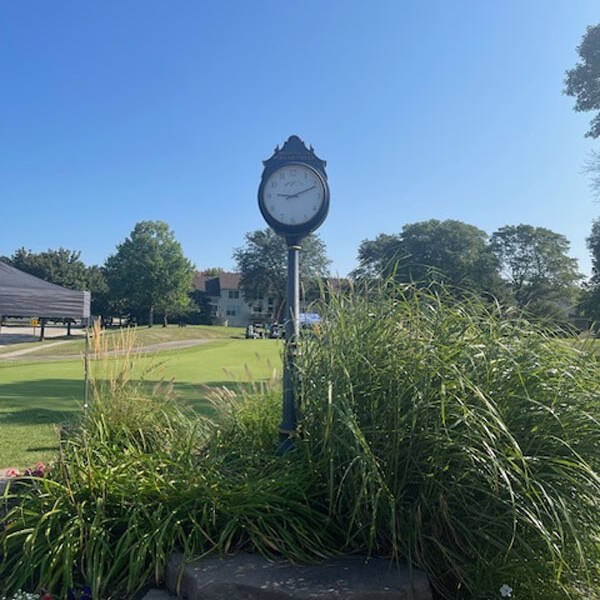 A vintage clock stands amid lush greenery and blooming flowers under a clear blue sky. A well-maintained green lawn, possibly perfect for a golf outing, and a building are visible in the background, suggesting a peaceful garden or park setting.