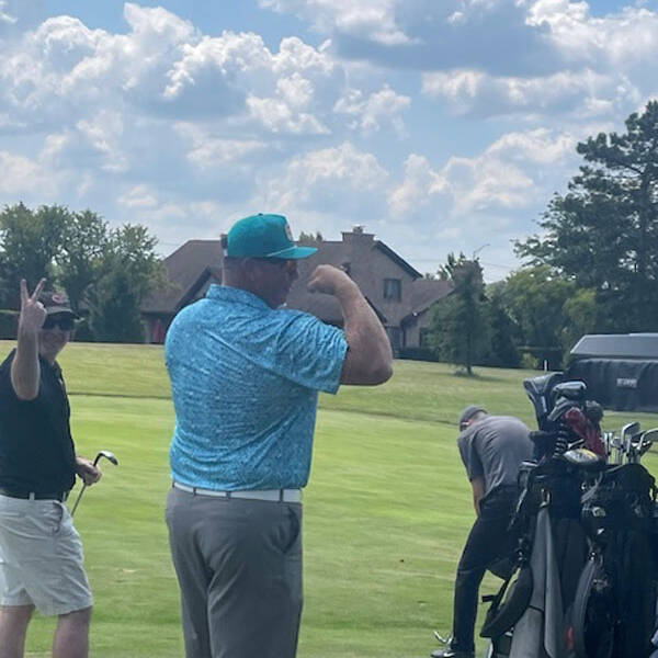 A man in a blue shirt and green cap flexes his muscles during the 2024 golf outing. Another person to his left makes a peace sign. In the background, a golfer leans over next to a golf cart filled with clubs, and a house and trees are visible under a cloudy sky.