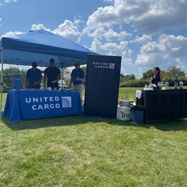 A grassy outdoor event setup with a blue canopy tent housing three people behind a table covered with a blue United Cargo tablecloth. To the right, a woman stands behind a mobile bar with various bottles and supplies. Set against a blue sky with clouds, this 2024 outing feels like the perfect day for golf.