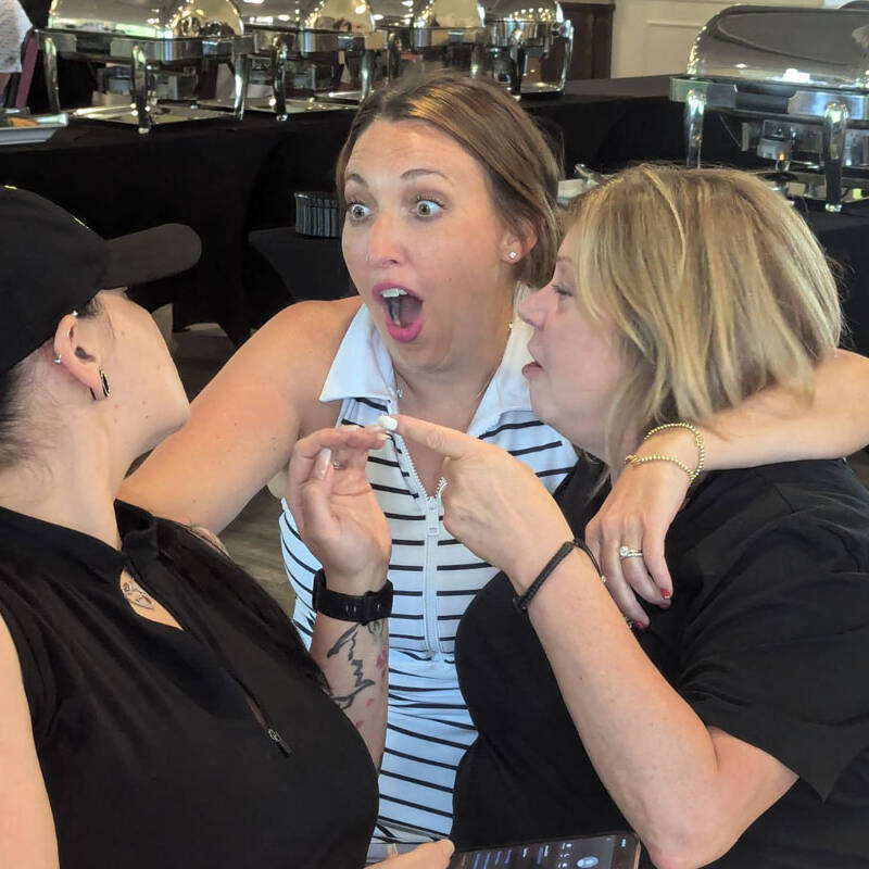 Three women are engaged in an animated conversation during an outing. The woman in the middle, appearing surprised, stands between one wearing a black cap and another holding a phone who is speaking intently. They're in a catering area with buffet trays visible in the background, perhaps enjoying a break from their 2024 golf event.