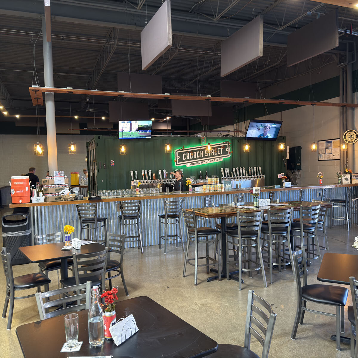 A modern brewery interior with a long bar counter featuring multiple beer taps invites you to the 2024 Oktoberfest. High tables and chairs are arranged in the foreground, surrounded by TV screens. Overhead lights illuminate a green "Church Street" sign above the bar, creating a festive atmosphere.