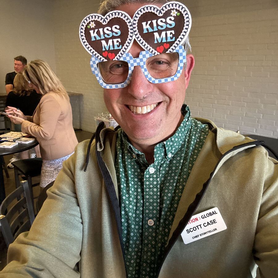 A person wearing novelty glasses with heart-shaped frames that say "Kiss Me" smiles at the camera during an Oktoberfest event. They are in a room with a few people in the background, sporting a green shirt and beige jacket.