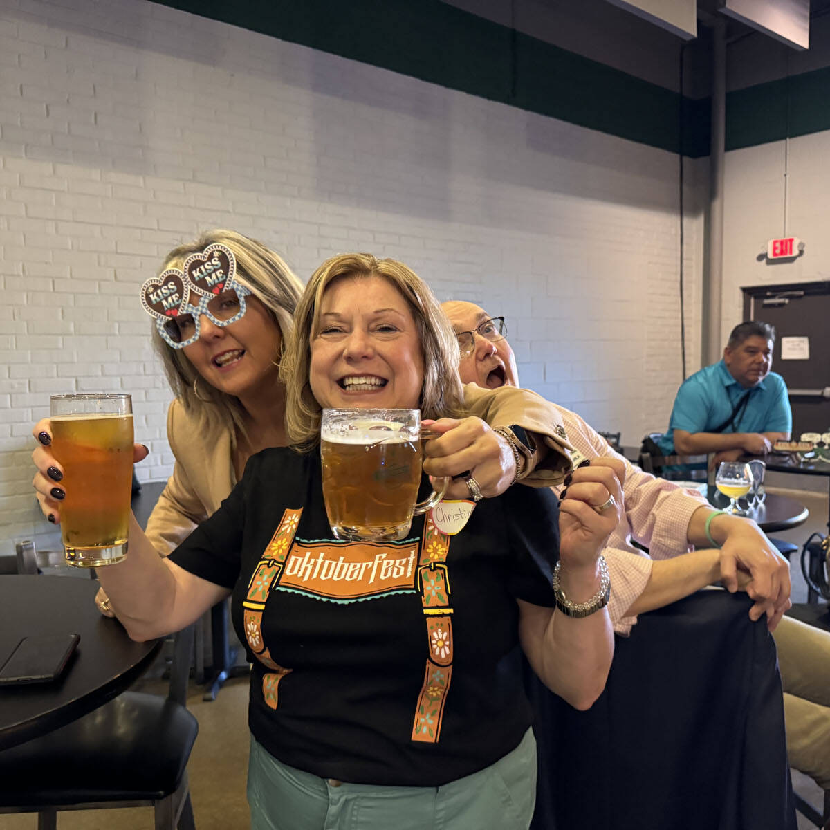 At a lively Oktoberfest event, three people enjoy drinks at a casual venue. The woman in front holds a beer, smiling widely, while the woman beside her sports novelty heart-shaped glasses. A man in the background playfully leans in, surrounded by tables of cheerful patrons.