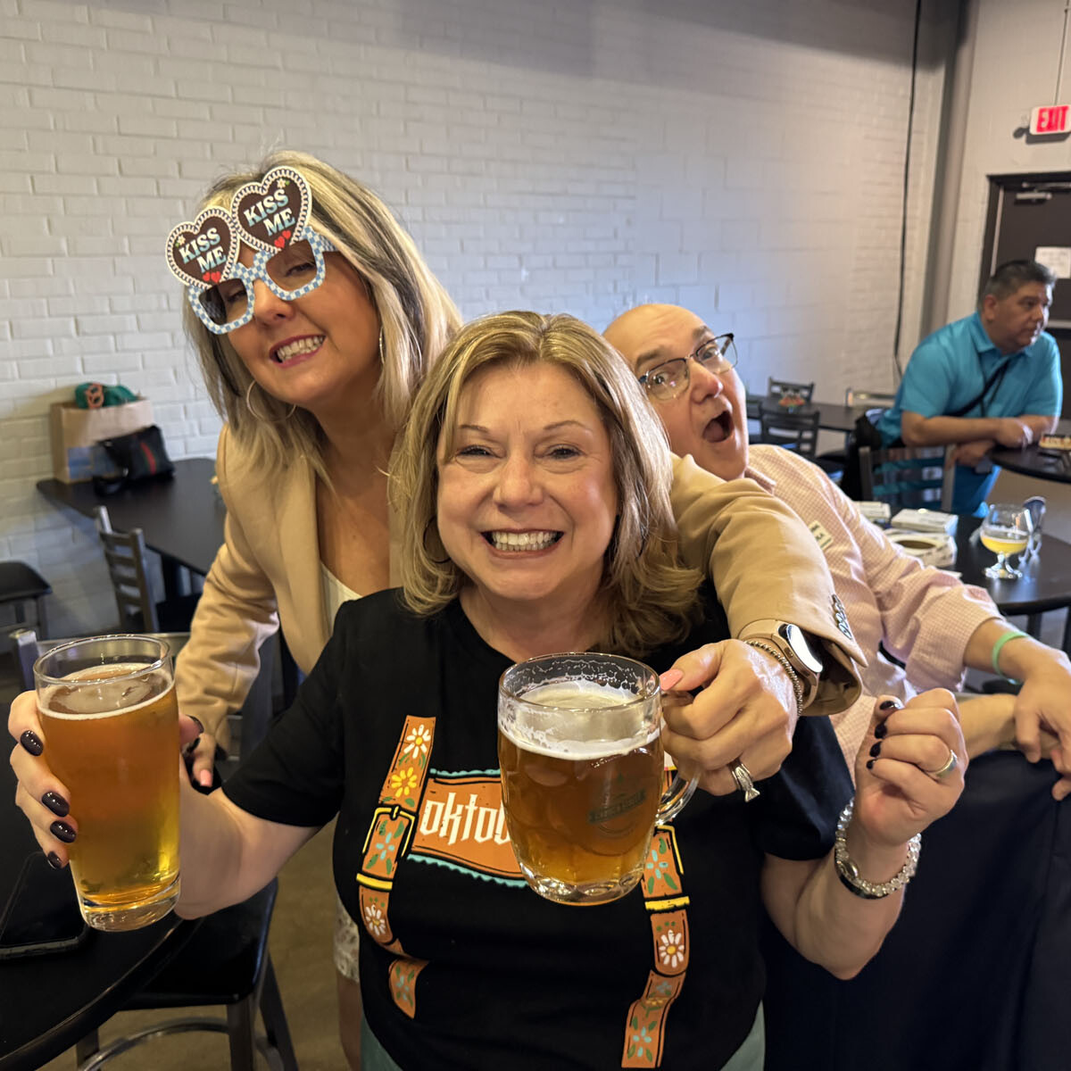 Three people enjoying beers at an Oktoberfest 2024 event. A woman in heart-shaped glasses leans in playfully from the left, while the woman in the center grins, holding a beer. A man in the back makes a funny face. Others are seated in the background.