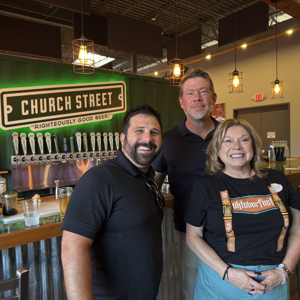 Three people are smiling inside a brewery, radiating the cozy, industrial vibes of an Oktoberfest celebration. Behind them, a row of beer taps with the sign "CHURCH STREET Righteously Good Beer!" enhances the festive mood. Hanging lights and a wooden bar counter complete this cheerful scene.