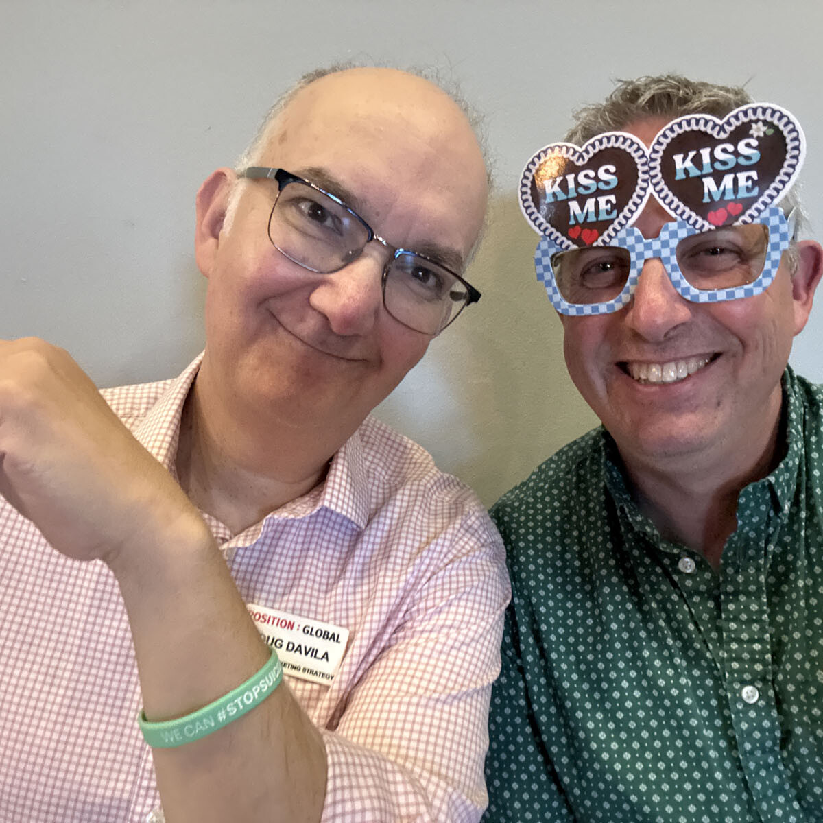 Two men grinning at the camera, one in a pink checkered shirt and glasses. The other sports a green button-up and heart-shaped novelty glasses with "KISS ME" boldly written. It's clear they're ready for some 2024 festival fun, perhaps even an Oktoberfest to remember!