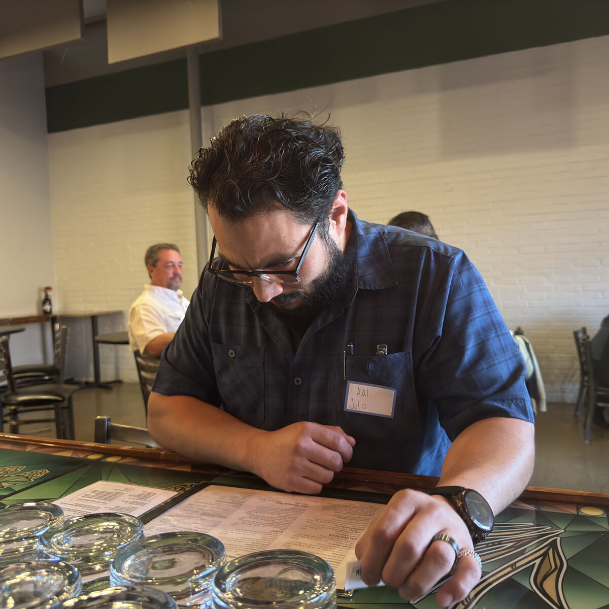 A man with dark hair and a beard is standing at a bar counter, wearing a blue plaid shirt with a name tag. At the Oktoberfest celebration, he leans over to read a menu, several clear glass bowls before him. Other festival-goers are seated in the background, enjoying the lively atmosphere.