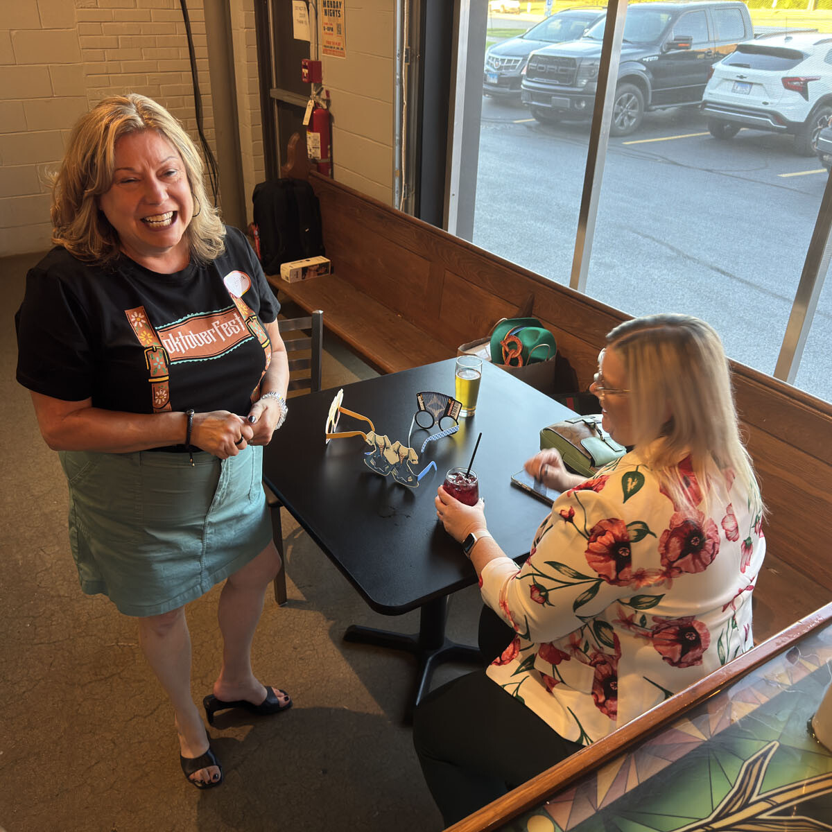 Two women are in a casual dining area. One stands smiling and wearing a black t-shirt, while the other sits at a table with drinks, wearing a floral blouse. They appear to be enjoying a friendly conversation, perhaps planning their next outing to Oktoberfest 2024.