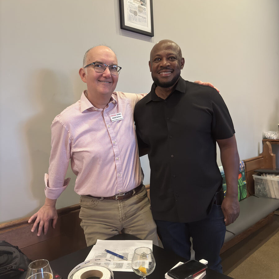 Two men are standing side by side, smiling at the camera. One is in a pink striped shirt, and the other wears black. They're in a room with a table holding papers, markers, and beverages. A framed document hangs on the wall behind them, ready for their 2024 festival planning session.