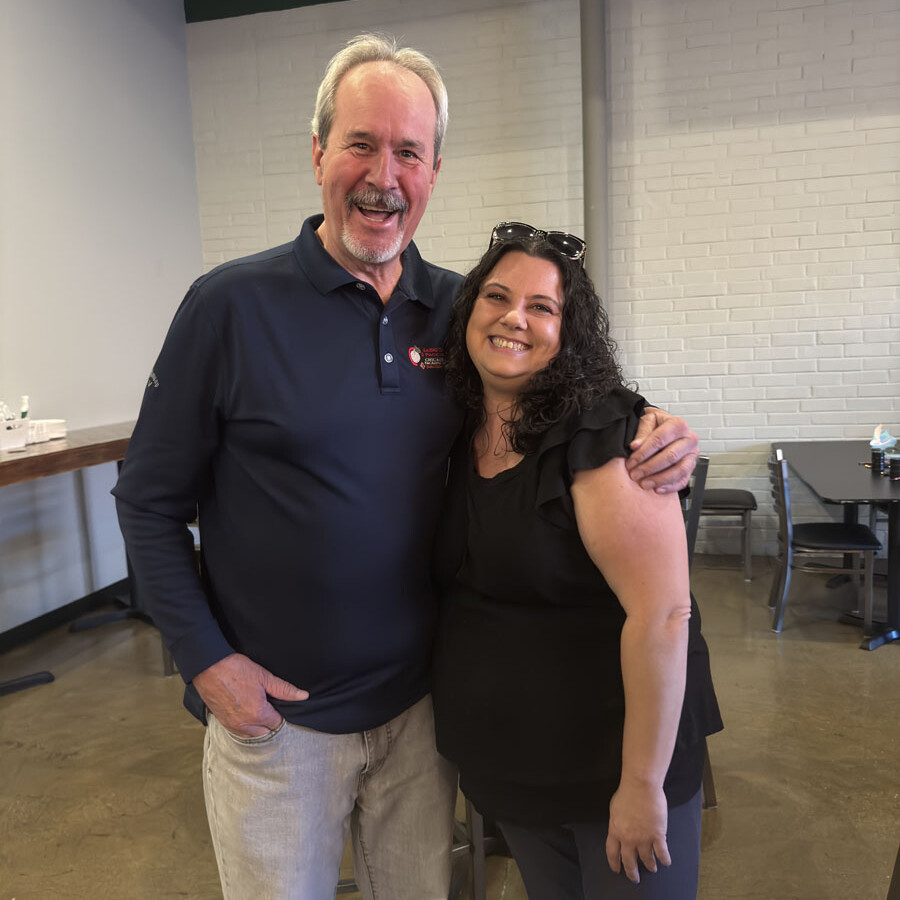 A smiling man and woman pose together indoors, both in casual attire, as if capturing a moment at an Oktoberfest festival. The bearded man wears a dark polo shirt with light pants, while the curly-haired woman dons a black blouse and pants. They stand against a white brick wall.