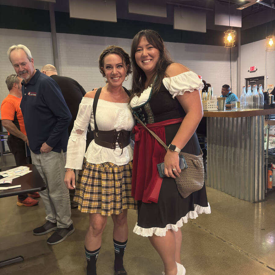 Two women in festive attire, one in a plaid skirt and the other in a black dress with red accents, capture the spirited essence of Oktoberfest indoors. They're smiling on a polished concrete floor, with people and tables bustling in the background.