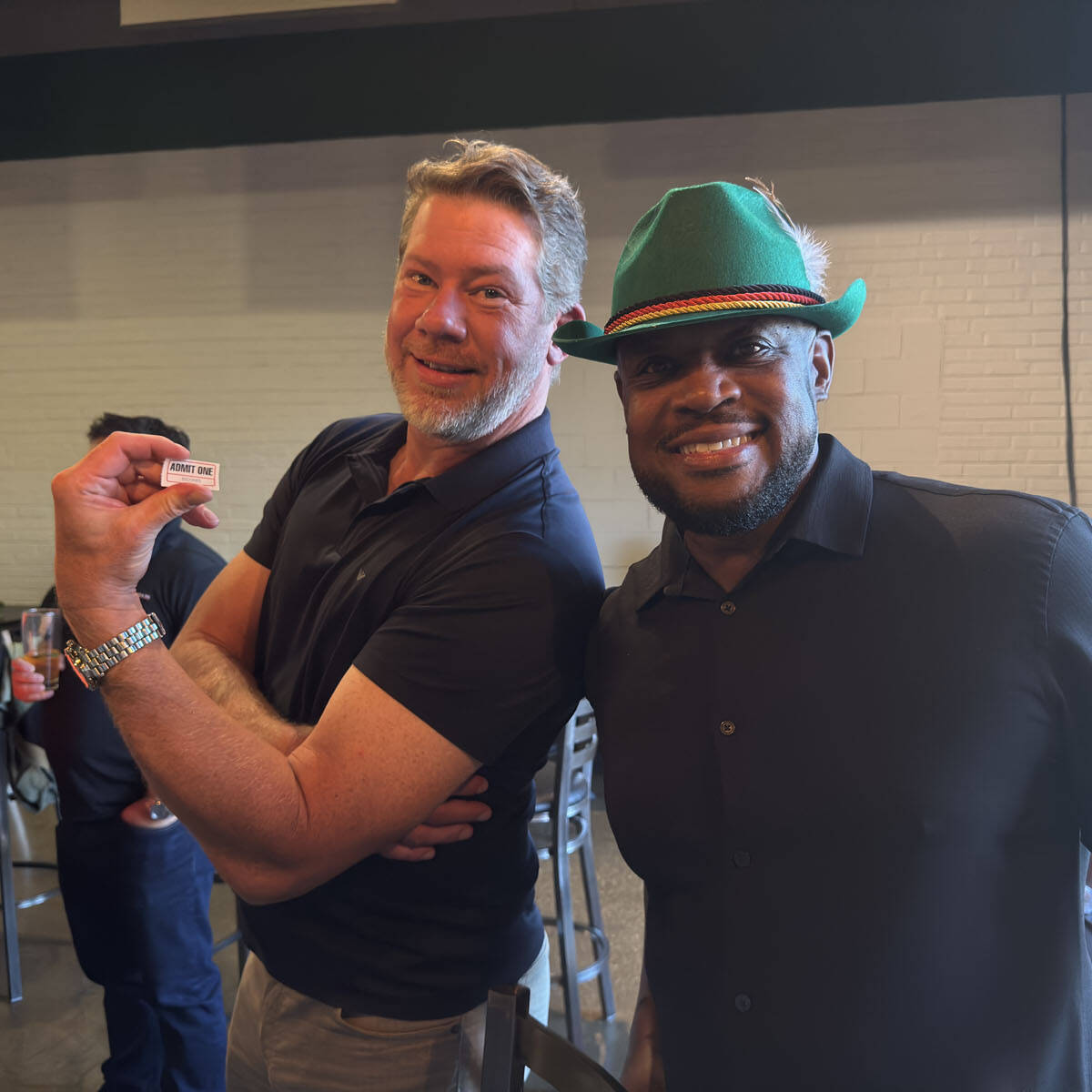 Two men smiling at the camera in a casual indoor setting, perhaps celebrating Oktoberfest. The man on the left holds a small rectangular card, while the man on the right sports a green hat with a red and yellow band. Happy festival-goers mingle in the background, capturing the spirit of 2024.