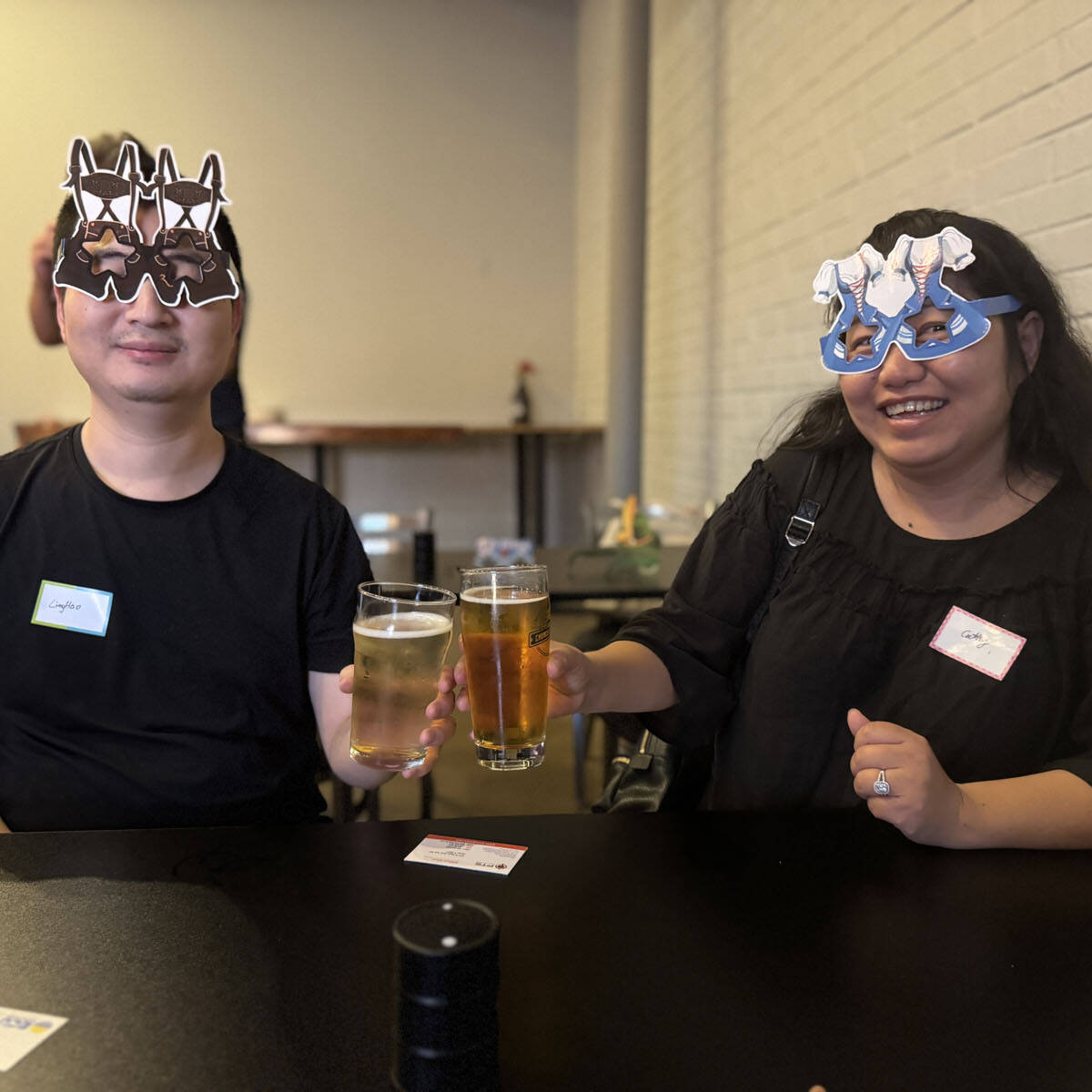 Two people wearing playful star-shaped masks hold glasses of beer, celebrating at an Oktoberfest event. They smile warmly, each sporting a name tag. The casual setting features a plain white brick wall with tables and chairs in the background.