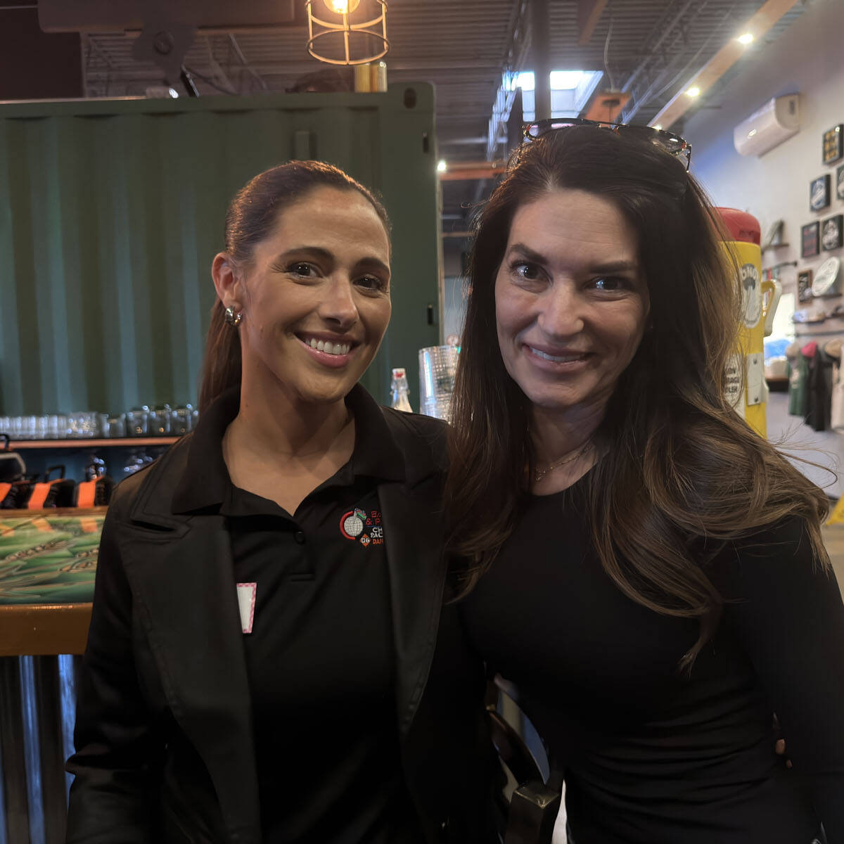 Two women smiling and standing together in an indoor setting, capturing the lively spirit of Oktoberfest 2024. One wears a black jacket with a nametag; the other dons a black long-sleeve top. Behind them, a bar counter with taps and decorated walls adds to the festive atmosphere.